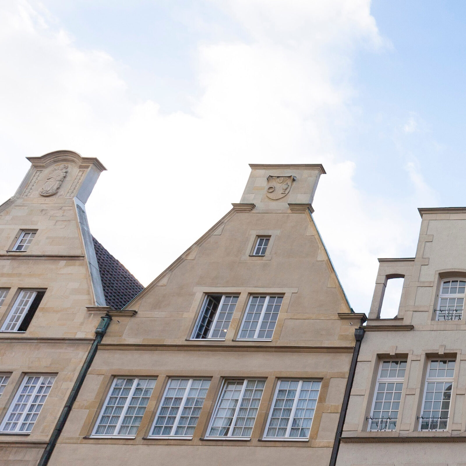 Leicht seitliche Ansicht mit Blick von unten auf eine alte braune Haeuserfront mit vielen Fenstern. Darueber ist der blaue Himmel mit weißen Wolken zu sehen.