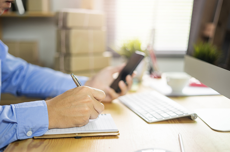 Haende einer Person mit blauem Hemd, die an einem Holzschreibtisch sitzt. In der linken Hand haelt sie ein schwarzes Handy, mit der rechten Hand schreibt sie in ein Notizbuch. Auf dem Tisch stehen zudem eine weiße Tastatur, ein Computerbildschirm und im Hintergrund eine weiße Tasse sowie etwas Deko.