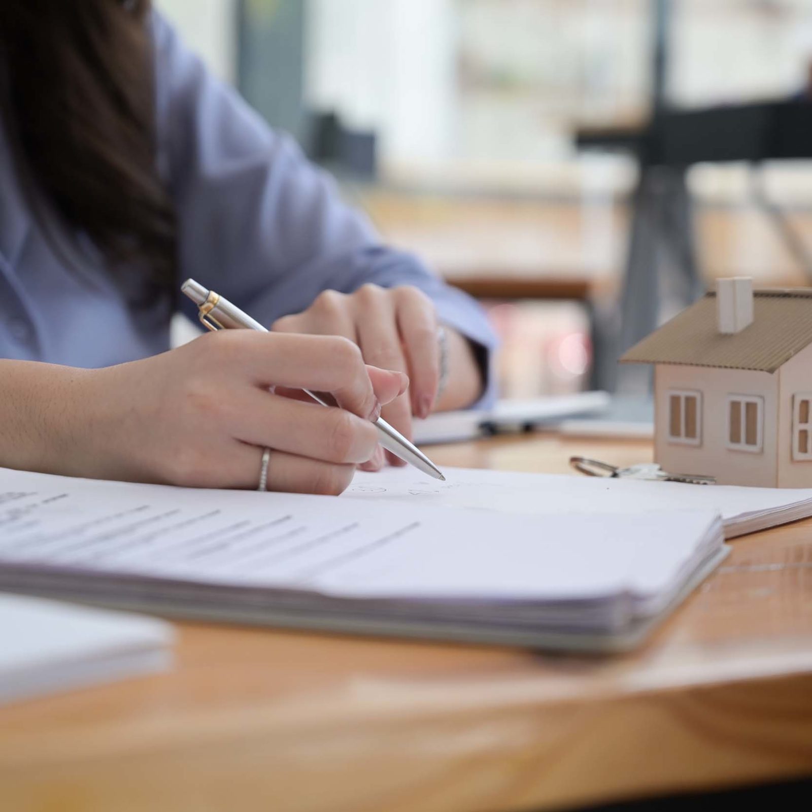 Frau mit langen braunen Haaren und blauem Oberteil sitzt an einem Holztisch. In der einen Hand hat sie einen Stift und deutet auf ein Dokument, das auf einem Stapel liegt. Daneben liegen weitere Dokumentenstapel. Ihr gegenueber sitzt eine weitere Person, die die Hand auf dem Tisch abgelegt hat. Auf dem Tisch steht ein kleines Dekohaus mit weißen Waenden und braunem Dach.