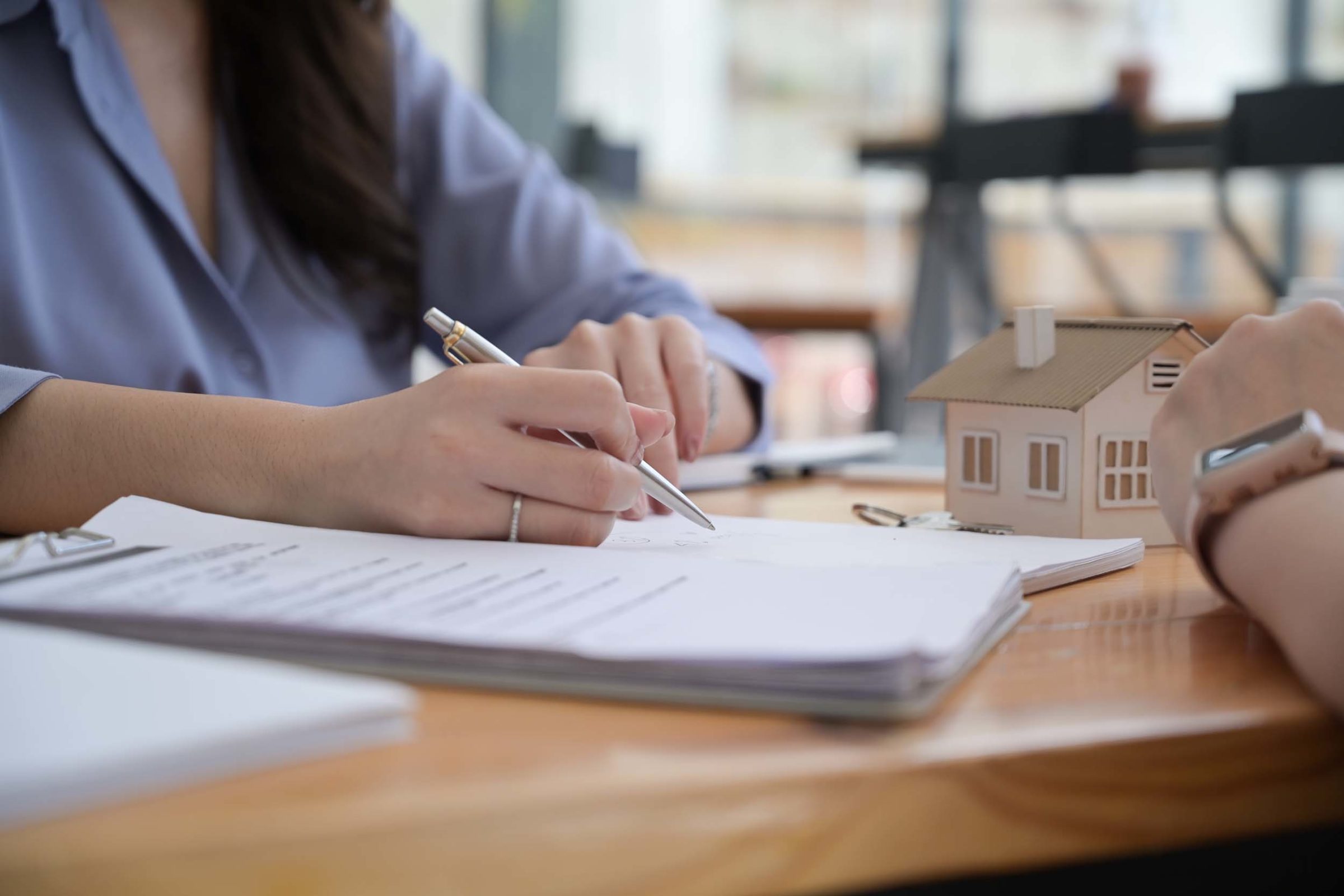 Frau mit langen braunen Haaren und blauem Oberteil sitzt an einem Holztisch. In der einen Hand hat sie einen Stift und deutet auf ein Dokument, das auf einem Stapel liegt. Daneben liegen weitere Dokumentenstapel. Ihr gegenueber sitzt eine weitere Person, die die Hand auf dem Tisch abgelegt hat. Auf dem Tisch steht ein kleines Dekohaus mit weißen Waenden und braunem Dach.