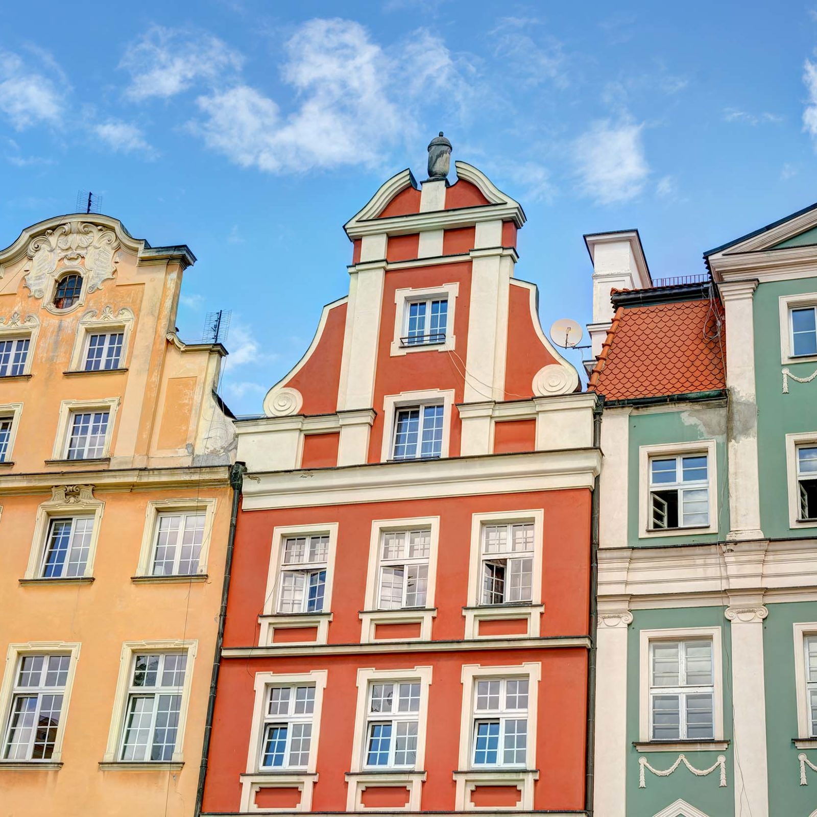 Denkmalschutz bei Frontansicht einer bunten Haeuserreihe in geld, rot und gruen mit unterschiedlichen Daechern, vielen Fenstern und Stuckverzierungen. Der Himmel ist blau mit wenigen weissen Wolken.