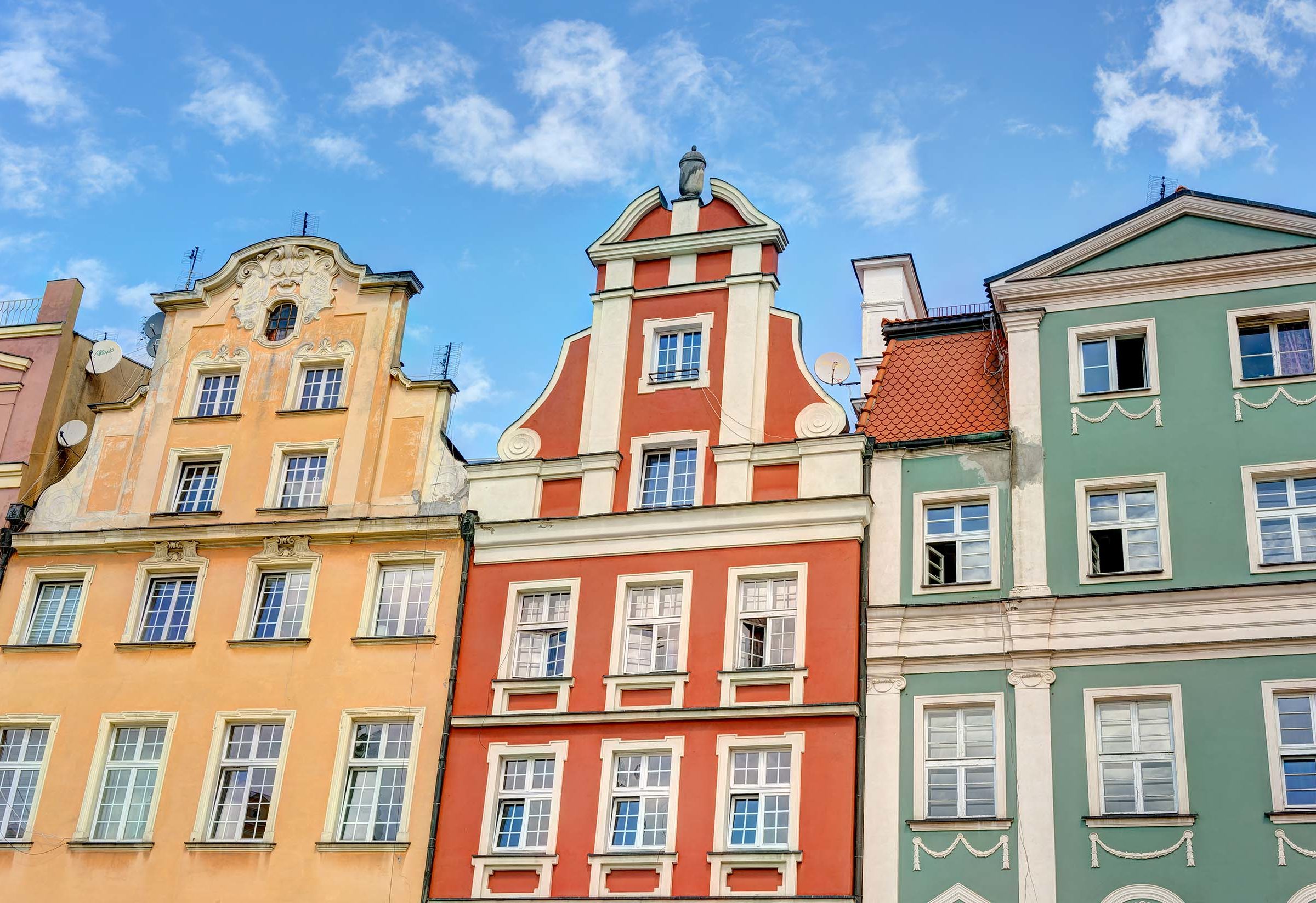 Denkmalschutz bei Frontansicht einer bunten Haeuserreihe in geld, rot und gruen mit unterschiedlichen Daechern, vielen Fenstern und Stuckverzierungen. Der Himmel ist blau mit wenigen weissen Wolken.