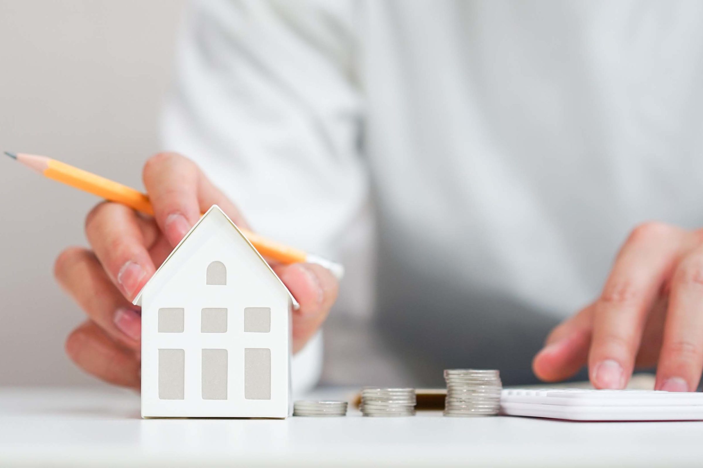 Haende einer Person mit hellem Oberteil, die hinter einem weißen Tisch sitzt. Vor ihr stehen ein kleines weißes Dekohaus, mehrere kleine Muenzstapel und ein Taschenrechner. Die eine Hand haelt einen Bleistift fest und hat drei Finger auf dem Dach des Hauses abgelegt. Die andere Hand bedient mit dem Zeigefinger den Taschenrechner.