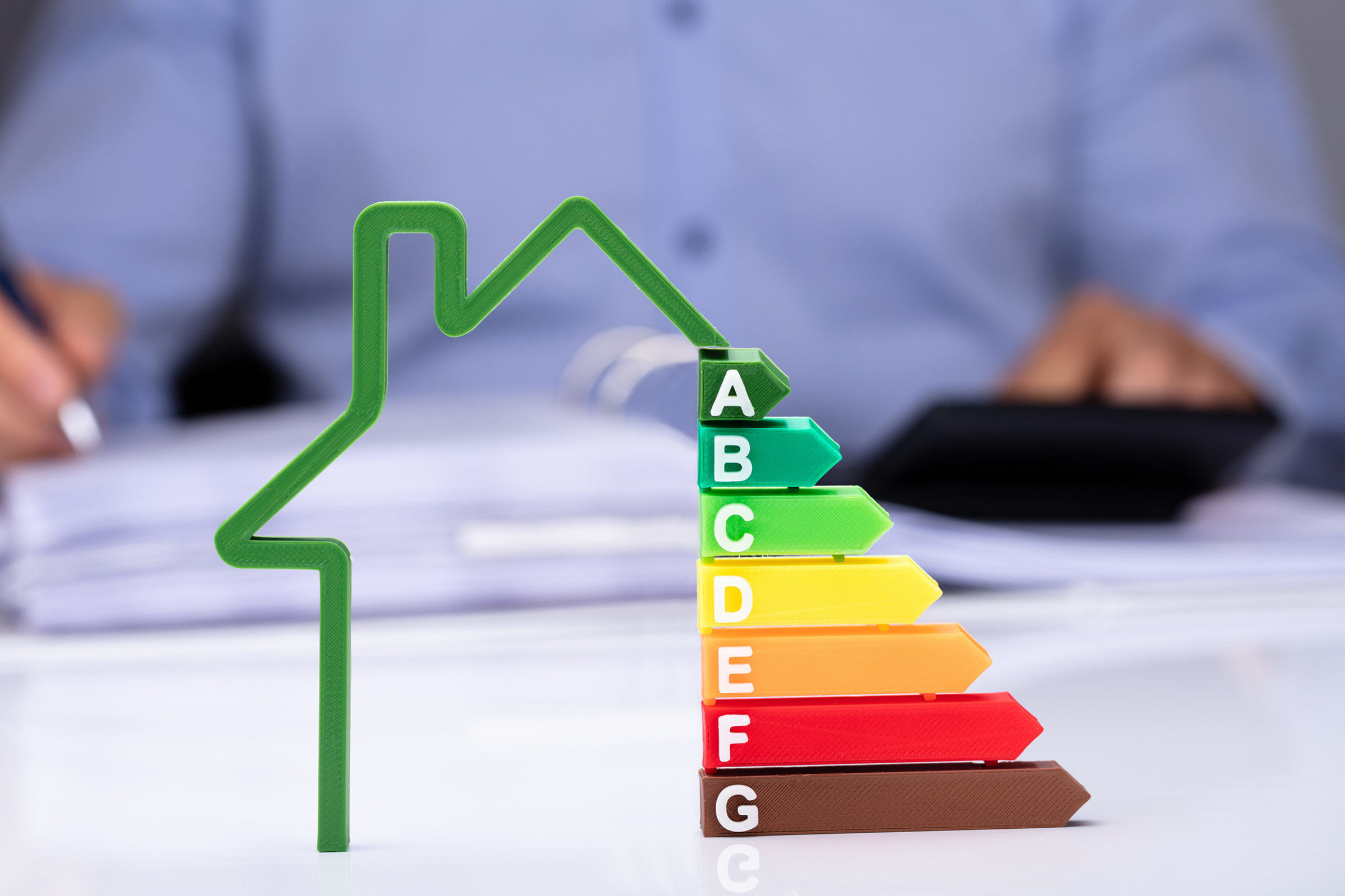 Stilisiertes gruenes Haus, auf der rechten Seite sind die Energieeffizienzklassen als bunte Elemente eingefuegt. Im Hintergrund sieht man den Oberkoerper eines Mannes mit blauem Hemd und auf einem Tisch liegen ein Taschenrechner und Dokumente, mit denen der Mann arbeitet.