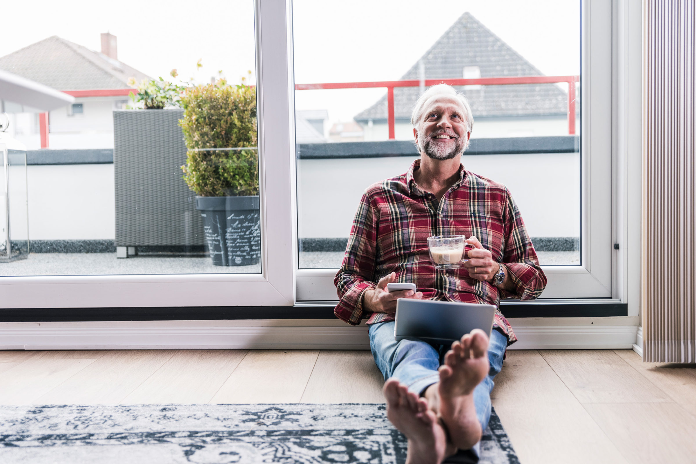 Mann mit kariertem Hemd sitzt auf dem Boden vor einem grossen Fenster. Er haelt eine Tasse in der Hand, auf seinem Schoss liegt ein Laptop und er streckt seine Fuesse Richtung Kamera. Auf dem Boden liegt ein großer Teppich. Auf dem Balkon hinter ihm stehen zwei Pflanzkuebel.