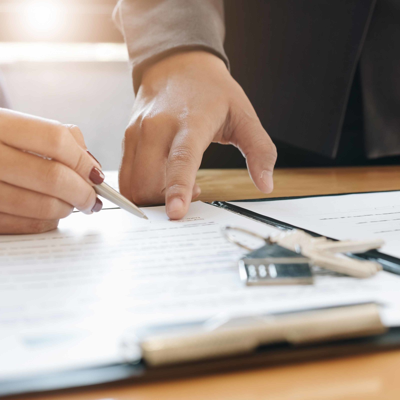 Eine Person unterschreibt ein Dokument. Eine andere Person deutet auf die passende Stelle. Auf dem Dokument liegt ein Schluesselbund mit einem Haus als Anhaenger.