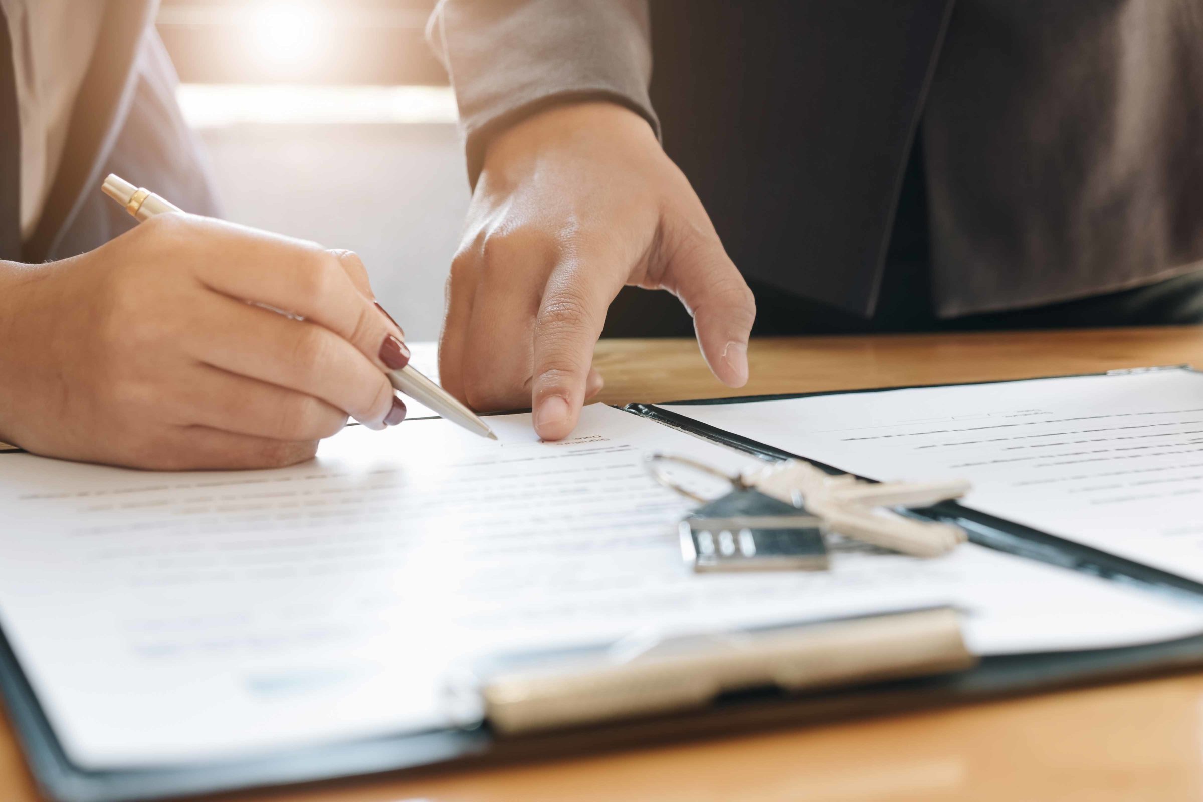 Eine Person unterschreibt ein Dokument. Eine andere Person deutet auf die passende Stelle. Auf dem Dokument liegt ein Schluesselbund mit einem Haus als Anhaenger.