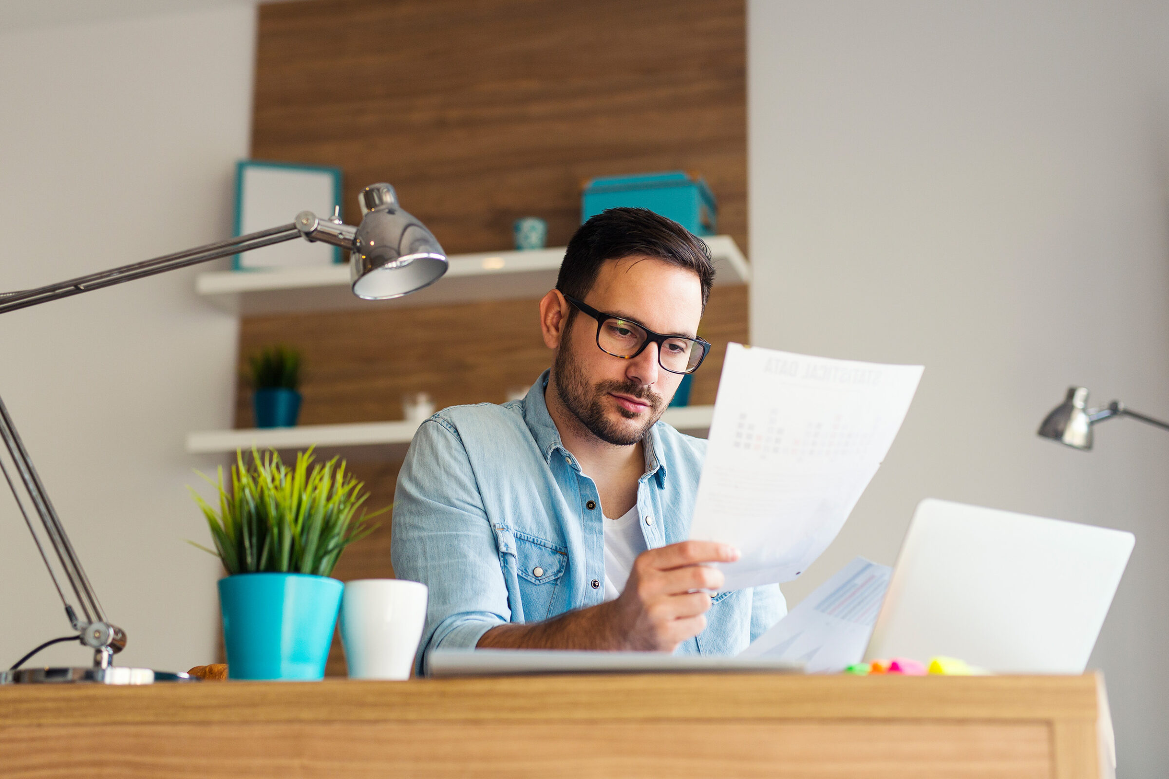 Widerspruch gegen Heizkostenabrechnung: In der Mitte des Bilds sitzt ein Mann mit dunklen Haaren, Bart und Brille mittleren Alters an einem Schreibtisch. Man sieht einen Teil des Oberkoerpers des Mannes, der ein blaues Jeanshemd und weißes T-Shirt traegt. In der rechten Hand haelt er ein Dokument, auf das er seinen Blick richtet. Vor ihm steht auf der rechten Bildseite ein weisser und aufgeklappter Laptop und es sind weitere Dokumente zu sehen. Ganz links im Bild ist im Hintergrund eine silberne Stehlampe zu sehen. Auf der linken Bildseite steht ebenfalls eine silberne Lampe auf dem braunen Schreibtisch aus hellem Holz. Im Vordergrund neben der Lampe stehen ein kleiner tuerkiser Blumentopf mit einer gruenen Pflanze und ein kleinerer weisser und leerer Blumentopf. Im Hintergrund des Bilds sieht man eine groesstenteils weisse Wand, mit einem Balken aus Holz. An diesem sind zwei weisse Regalbretter angebracht, auf denen blaue Kisten, Blumentoepfe und ein Bilderrahmen stehen.